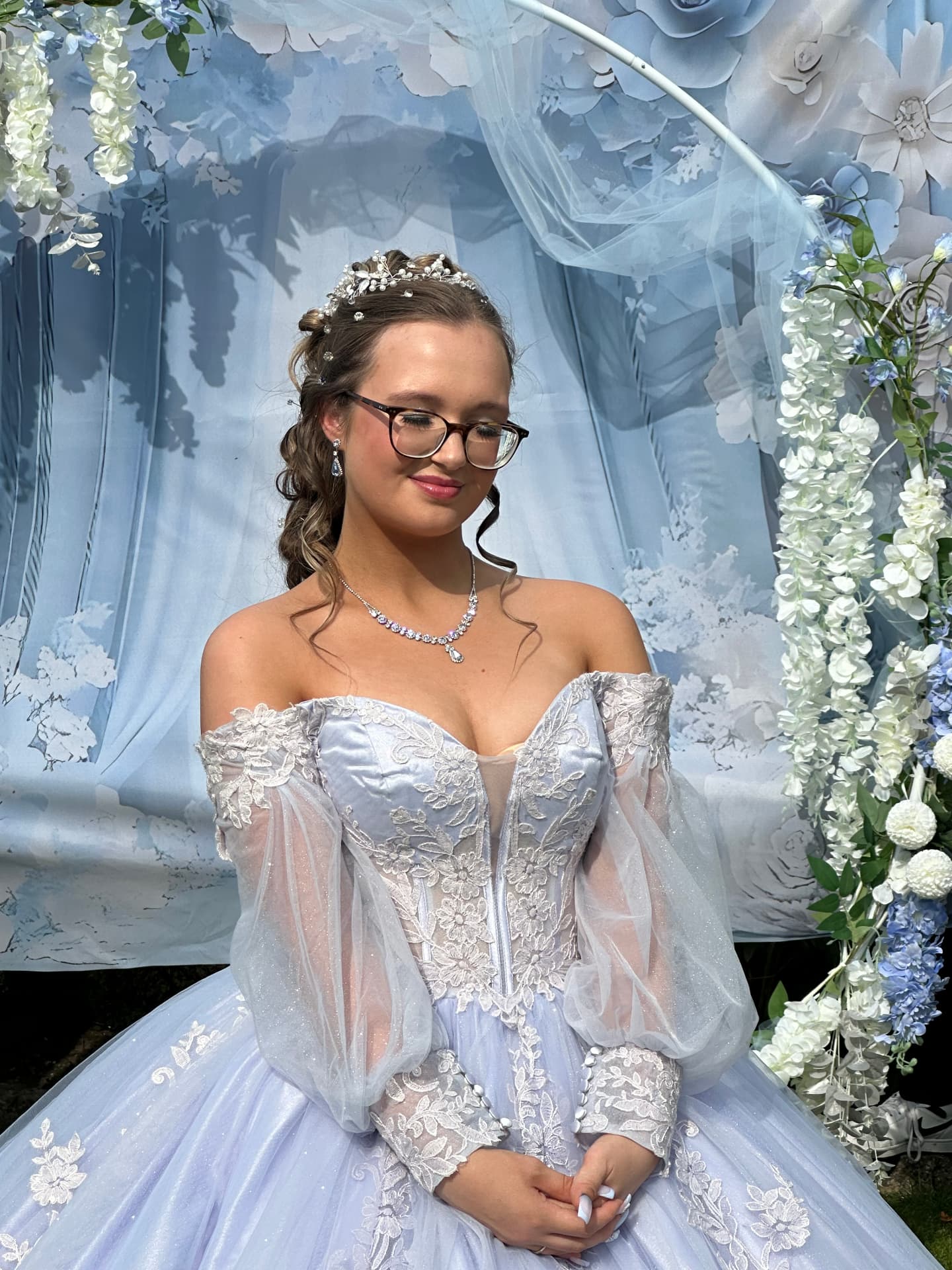Prom client in a blue gown with glam makeup by a floral arch